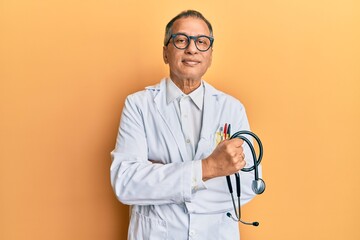 Middle age indian man wearing doctor coat holding stethoscope relaxed with serious expression on face. simple and natural looking at the camera.