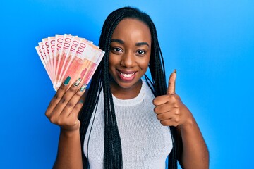 Young african american woman holding 100 norwegian krone banknotes smiling happy and positive,...