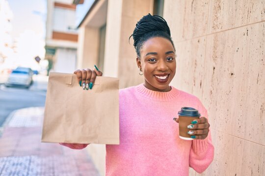 Young african american woman holding delivery paper bag and take away coffee at the city.