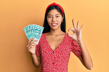 Young chinese woman holding 50 yuan chinese banknotes doing ok sign with fingers, smiling friendly gesturing excellent symbol - obrazy, fototapety, plakaty