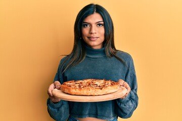 Young latin transsexual transgender woman holding italian pizza relaxed with serious expression on face. simple and natural looking at the camera.