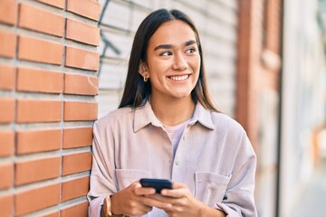 Young latin girl smiling happy using smartphone at the city.