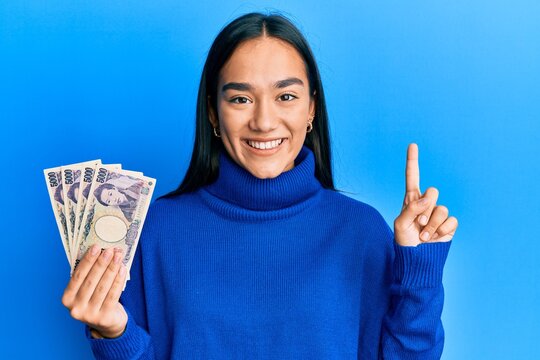Young asian woman holding 5000 japanese yen banknotes smiling with an idea or question pointing finger with happy face, number one