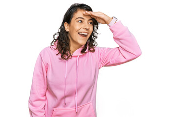 Young hispanic woman wearing casual sweatshirt very happy and smiling looking far away with hand over head. searching concept.