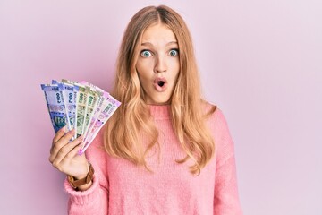 Beautiful young caucasian girl holding indian rupee banknotes scared and amazed with open mouth for...