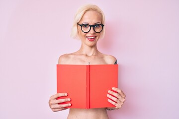 Young blonde woman with tattoo standing shirtless reading book smiling and laughing hard out loud because funny crazy joke.