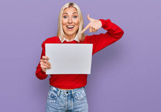Young blonde woman holding blank empty banner smiling happy pointing with hand and finger