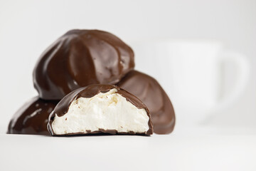 A macro photo of a half marshmallow covered with natural chocolate in the foreground. Medium shot with a group of other marshmallows and the background with a white cup is blurred.