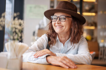 Beautiful mature senior woman in glasses and a hat sitting in a cafe and smiling looking at the...