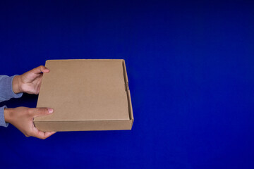 hands of a person holding a closed box from the left side, close up and good view of the top of the box, ideal for branding, giving a sense of delivering something, blue background and space for text