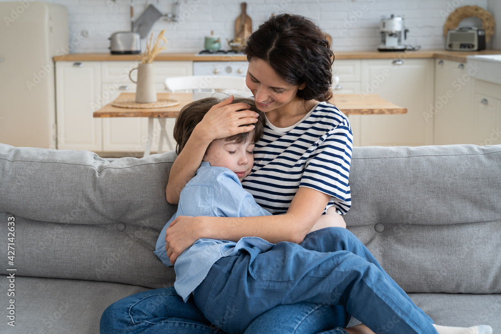 Wall mural head shot of happy young mom sit on sofa embrace tired after funny games at home son sleeping on her