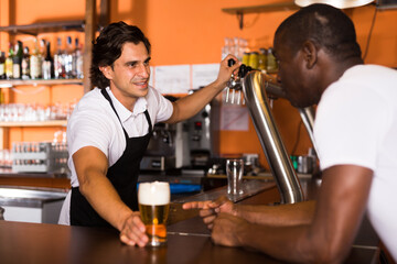 Smiling man barman giving glass of golden beer to client in cafe