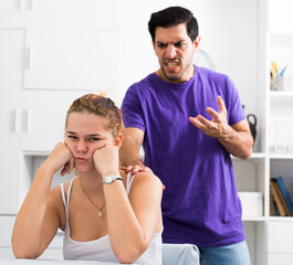 Frustrated girl sitting at home table on background with angry boyfriend