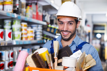 glad adult foreman standing near racks in paint store holding basket with tools