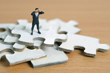Businessmen standing above jigsaw puzzle piece stack using binoculars above wooden table. Miniature tiny people toys photography.