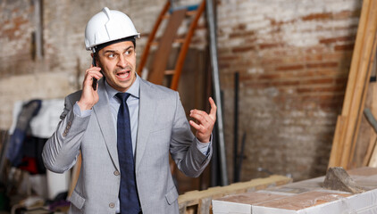 Portrait of angry manager in helmet talking on mobile phone on building