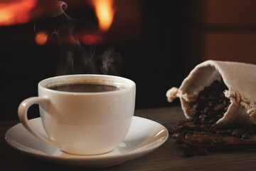 Close-up of cup of hot coffee and coffee beans in a bag on a wooden table in front of a burning fireplace. Selective focus