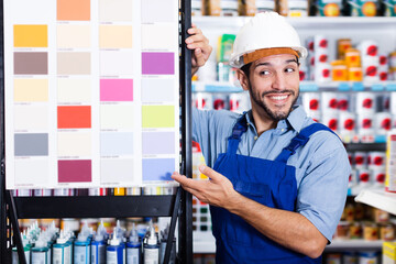 Cheerful foreman in blue overalls showing samples of paint on stand in store