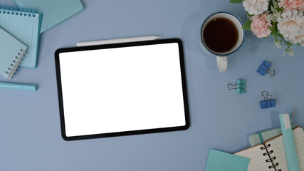 Top view of digital tablet with white screen, stylus pen, coffee cup and notebook on blue background.
