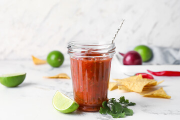 Glass jar of tasty salsa sauce with ingredients and nachos on light background
