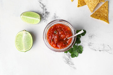 Glass jar of tasty salsa sauce with ingredients and nachos on light background