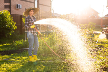 Caucasian woman gardener in work clothes watering the beds in her vegetable garden on sunny warm summer day. Concept of working in the garden and your farm