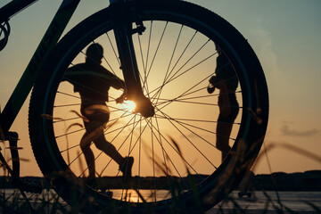 Naklejka premium Close-up silhouette of a bike wheel at sunset. The sun shines through the wheel of a bicycle with blurred runner figure at background