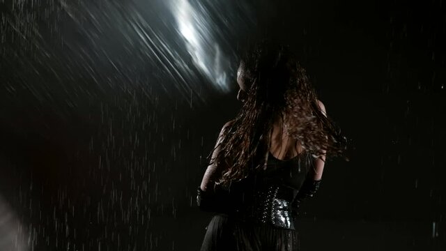 A hot young woman in a black sexy suit poses in a dark studio and in the rain. Water drops trickle down her body, she shakes her hair, slow motion.
