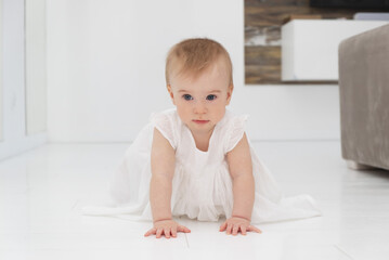 Fototapeta premium Cute little funny caucasian blonde baby girl in white dress, crawling on fours on floor at home