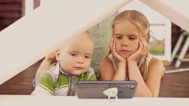 Two little blond-haired children closely looking in smartphone outdoors