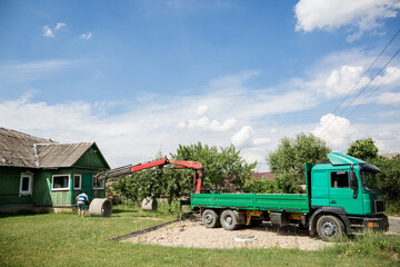 Obraz premium A truck with a crane manipulator unloads a concrete ring. Green lorry. Business transport cargo transportation. Delivery of bulky building material. Summer. Mobile crane