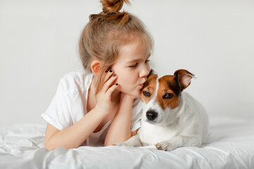 Beautiful funny kid girl lies in bed with a dog and kisses it.