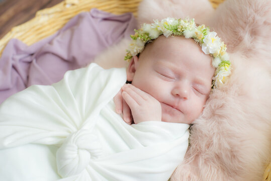 Baby girl in a wicker basket of vine decorated with  pink fur in a light winding and a flower wreath on her head. Spring photo. Flowers and children. Happy motherhood 