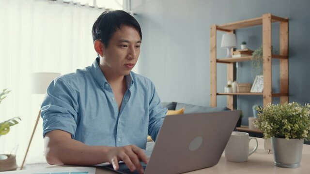 Freelance Asia guy casual wear using laptop online learning in living room at house. Working from home, remotely work, distance education, social distancing, quarantine for corona virus prevention.