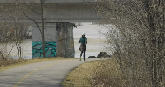 Woman runs down a bike path towards the rivers edge and a concrete bridge on the Ottawa River bike paths in Gatineau, Quebec. 