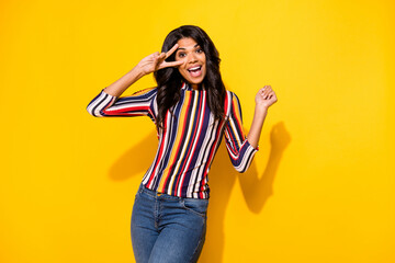 Portrait of adorable friendly dark skin lady beaming smile fingers show v-sign near eye isolated on yellow color background