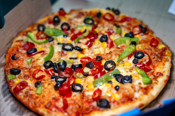 Selective focus of a vegetarian pan pizza topped with black olives, green bell peppers, red paprika, and golden corn in a takeout box