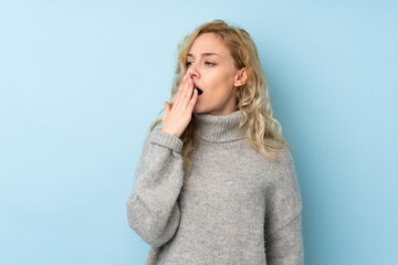 Young blonde woman wearing a sweater isolated on blue background yawning and covering wide open mouth with hand