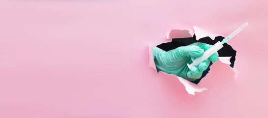 doctor's hand holds out a syringe with a vaccine through a hole in torn paper on a pink background