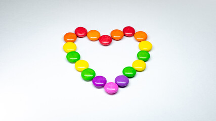 Selective focus of colorful button chocolates arranged in the shape of a heart on a white paper background.