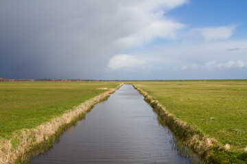 Fototapeta na wymiar Storm front approaching over rural landscape on the Dutch island Terschelling: dark threatening clouds above a flat polder landscape with a canal and dike at the horizon