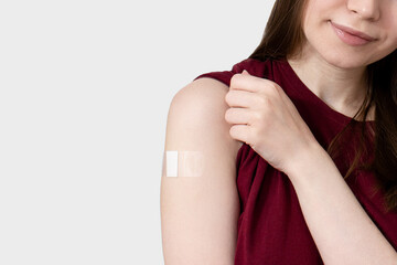 Smiling young woman in t-shirt showing plaster on shoulder after coronavirus vaccine, gray...