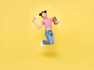 Happy Asian child girl jumping up with diploma and book isolated on yellow background.