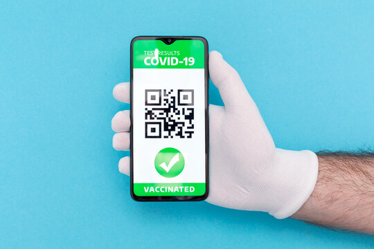 Concept of vaccination and digital passport. Hand in protective glove holding a cellphone with electronic Immunity passport with a COVID-19 vaccination stamp. Blue background. Flat lay