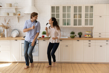 Overjoyed young married couple singing into kitchenware as microphone, playing, dancing in modern...
