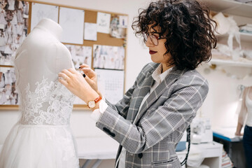 Work process of a tailor in her studio. Fashion designer sewing bridal dress, pinning lace on...