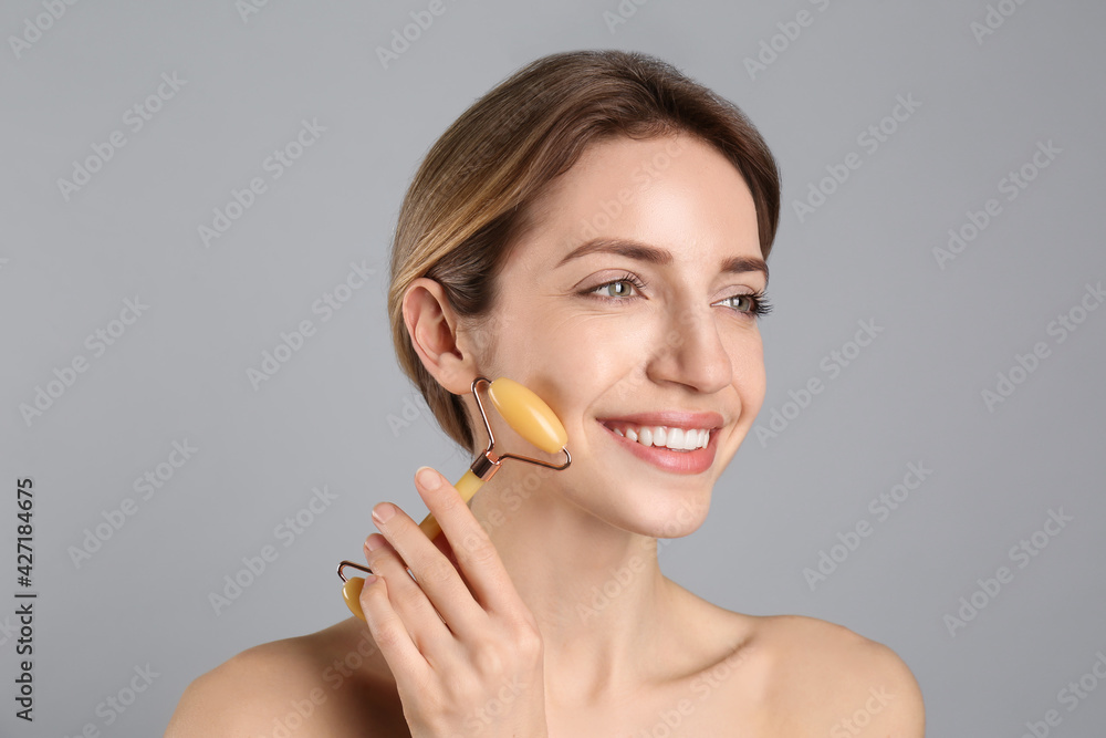 Poster Young woman using natural jade face roller on light grey background