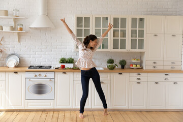 Overjoyed young woman tenant dancing, jumping in modern kitchen at home, excited by relocation in first own apartment, happy attractive young female having fun, moving to favorite music alone