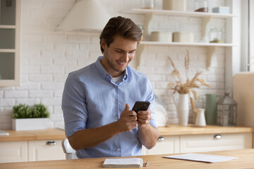 Smiling man holding smartphone, looking at screen, sitting at table in kitchen, having fun with...