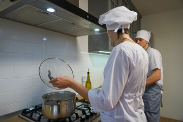 Two chefs in the restaurant kitchen prepare dinner. The chef is dressed in a white tunic and a chef's hat.